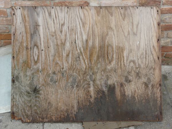 Light brown plywood texture, with a rough, scratched surface and a large dark stain at the bottom. Nails run along the edges.
